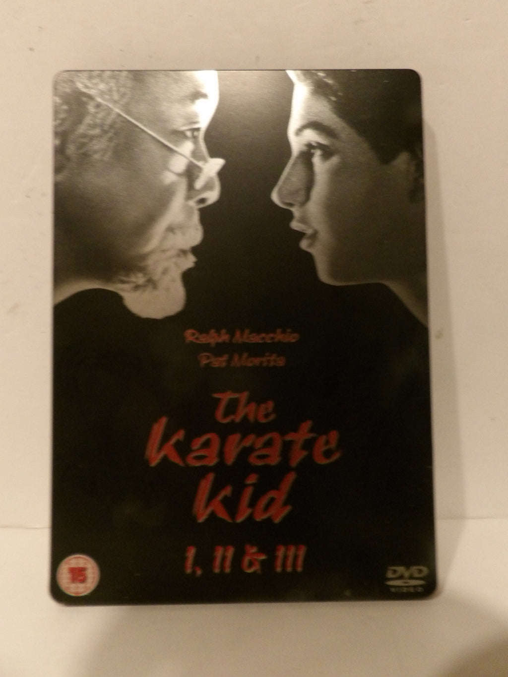 The Karate Kid 1, 11 & III DVD steelbook case on a light beige surface. The cover features a black and white graphic of Ralph Macchio and Pat Milla facing each other, with the title in red text.