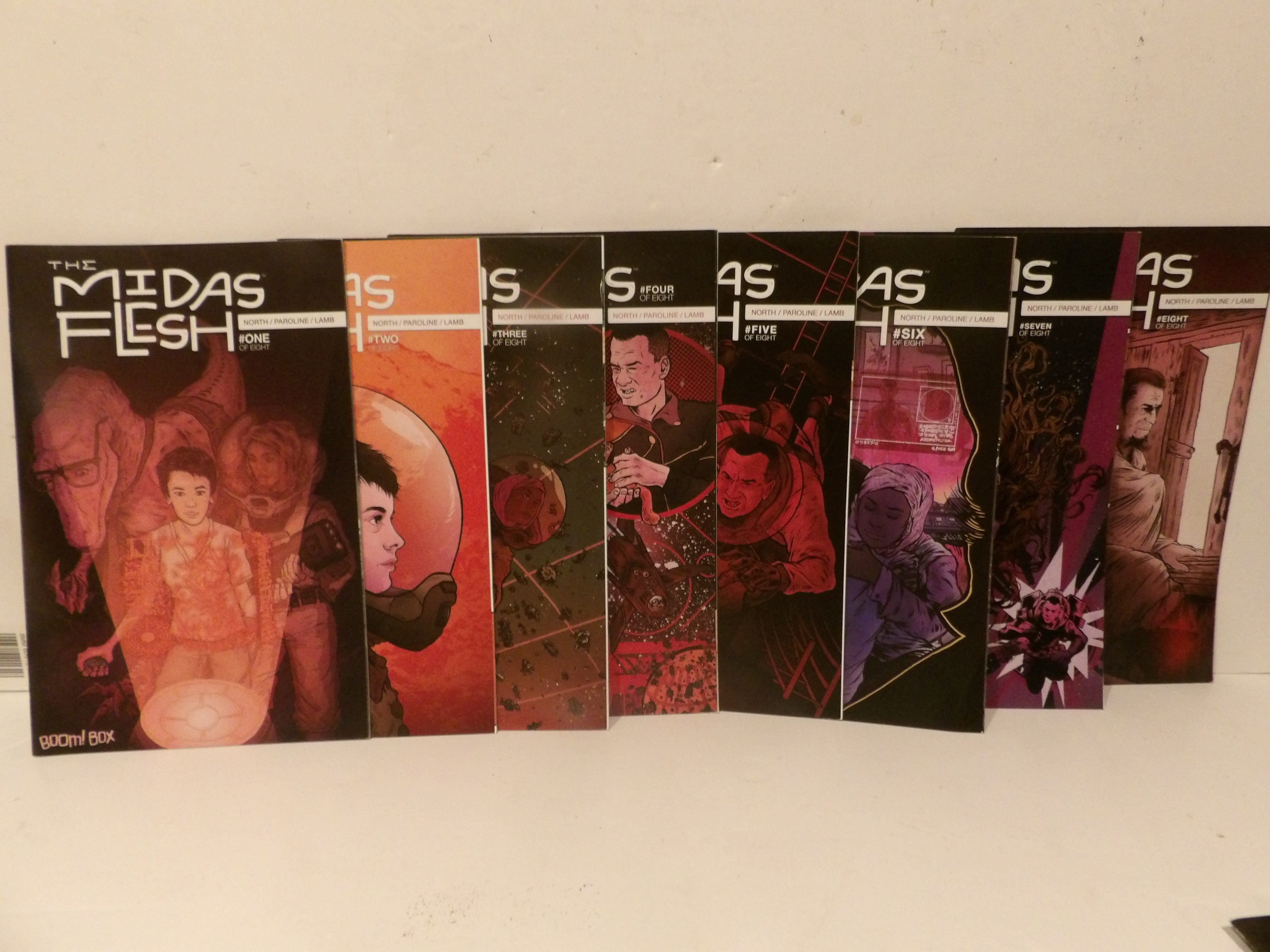 A row of seven comic book issues from The Midas Flesh Box arranged side-by-side on a light-colored surface against a plain wall. The covers feature various dark, sci-fi, and horror-themed illustrations in black, orange, red, and purple tones.