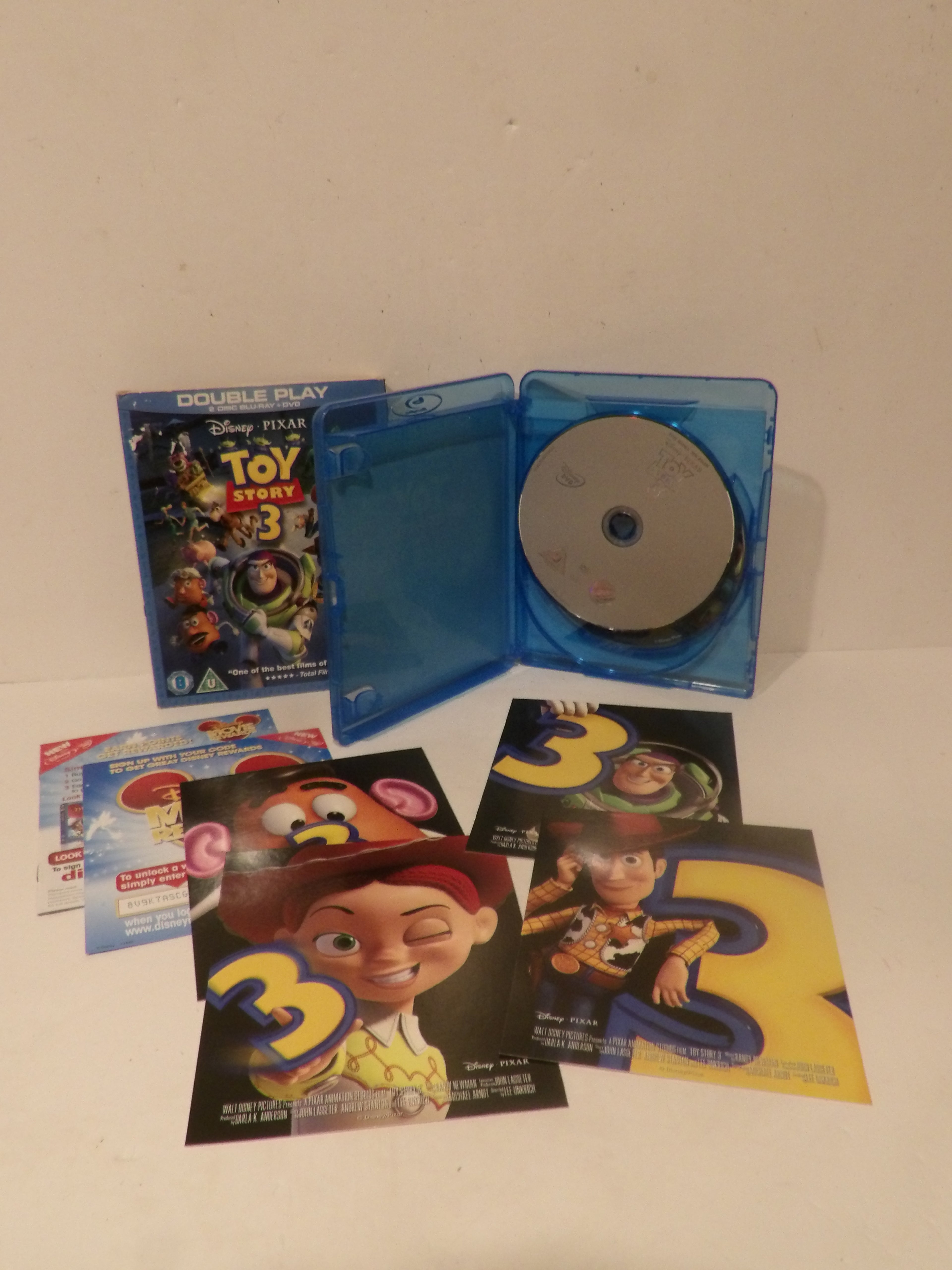 Toy Story 3 double play Blu-ray set with a blue plastic case open to show the disc, a Blu-ray case standing upright, and several character posters in front on a plain light background.