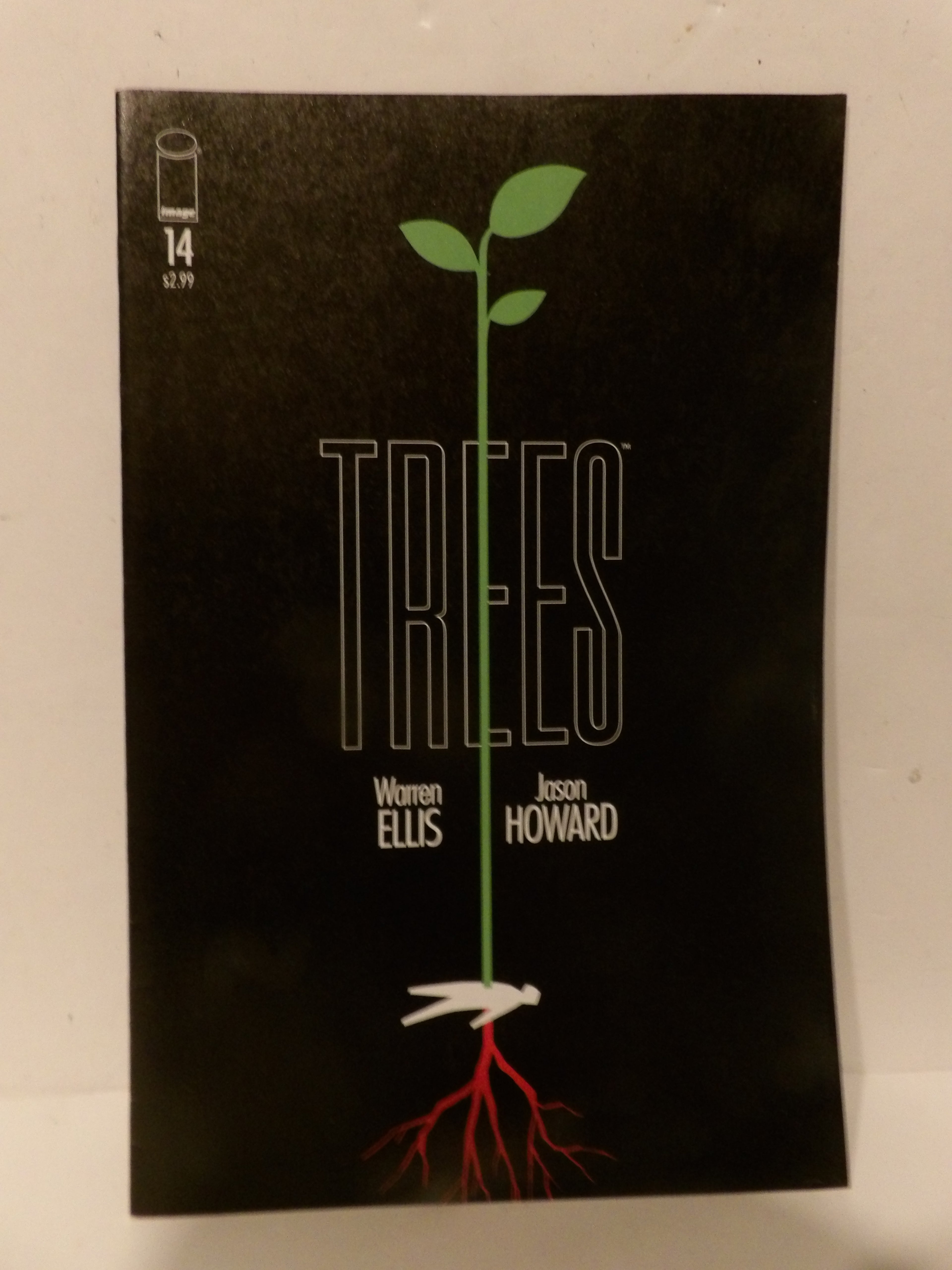 The front cover of the comic book Trees #14, featuring a black background with a stylized green sapling growing from a white bird, with red roots extending down. The title 'TREES' is written in large outlined letters, with creator names Warren Ellis and Jason Howard below.