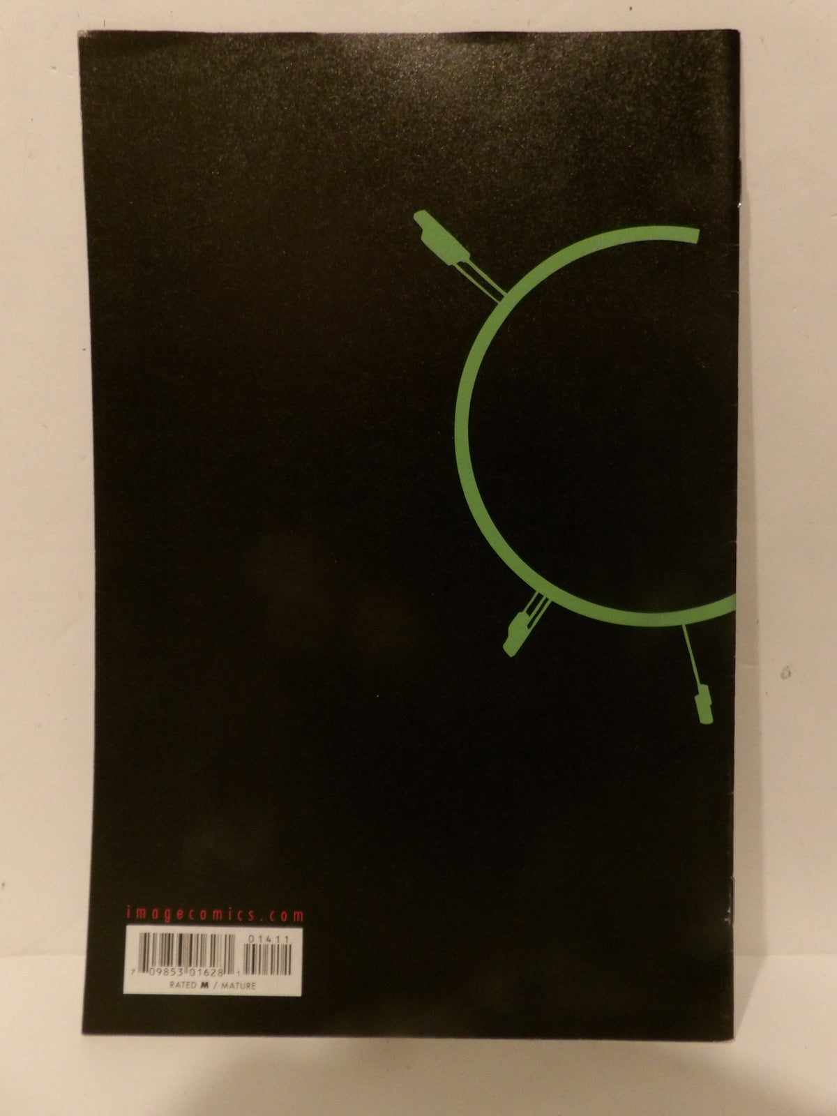 A black rectangular product, likely a comic book or notebook, featuring a green circular graphic and a barcode label at the bottom left, photographed against a light background.