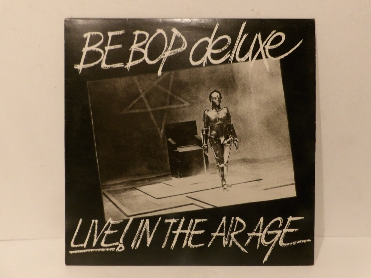 A square vinyl record sleeve for 'BE BOP deluxe LIVE! IN THE ARAGE' featuring a black and white photo of a performer on stage, photographed against a plain off-white background.