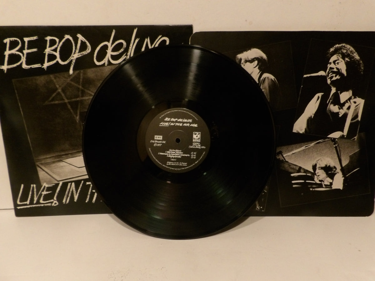 A black vinyl record partially pulled from its sleeve, with the album cover visible behind it. The cover has black-and-white photos of a performer and stylized white text reading 'BE BOP deli' and 'LIVE IN THE'.