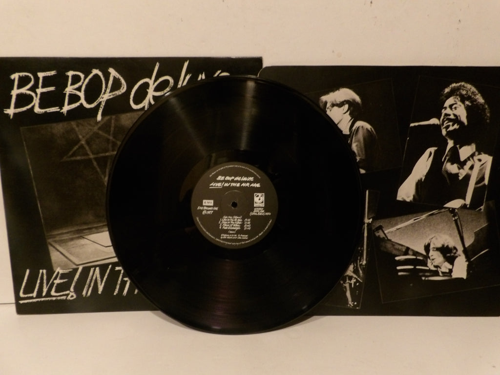 A black vinyl record partially covering the front cover of Be Bop Deluxe's 'Live! In Tokyo' album, with the album cover visible behind it on a light surface.