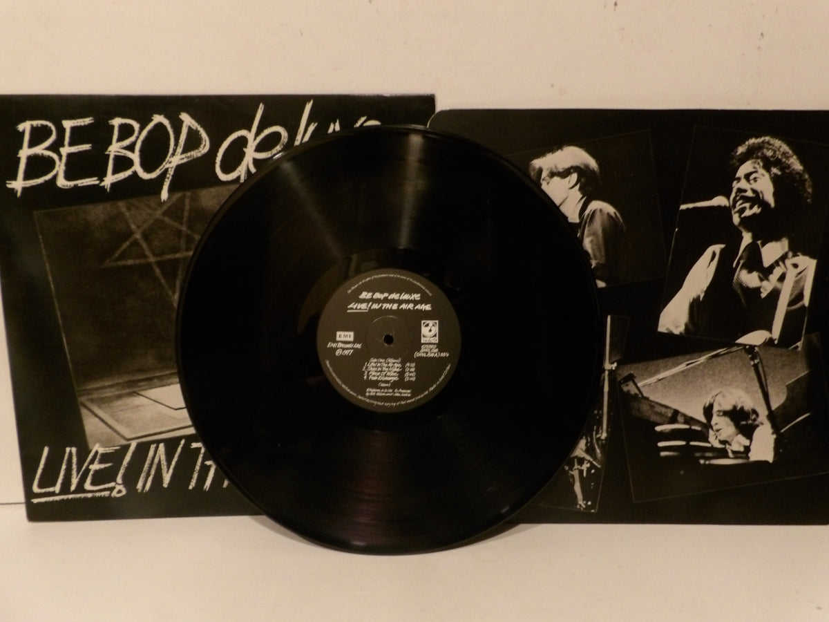 A black vinyl record partially covering the front cover of Be Bop Deluxe's 'Live! In Tokyo' album, with the album cover visible behind it on a light surface.